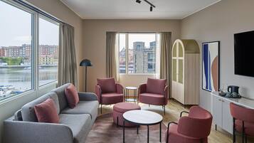 Een moderne hotelkamer met een grijze bank, rode fauteuils, een kleine ronde tafel en uitzicht op de stad door grote ramen.