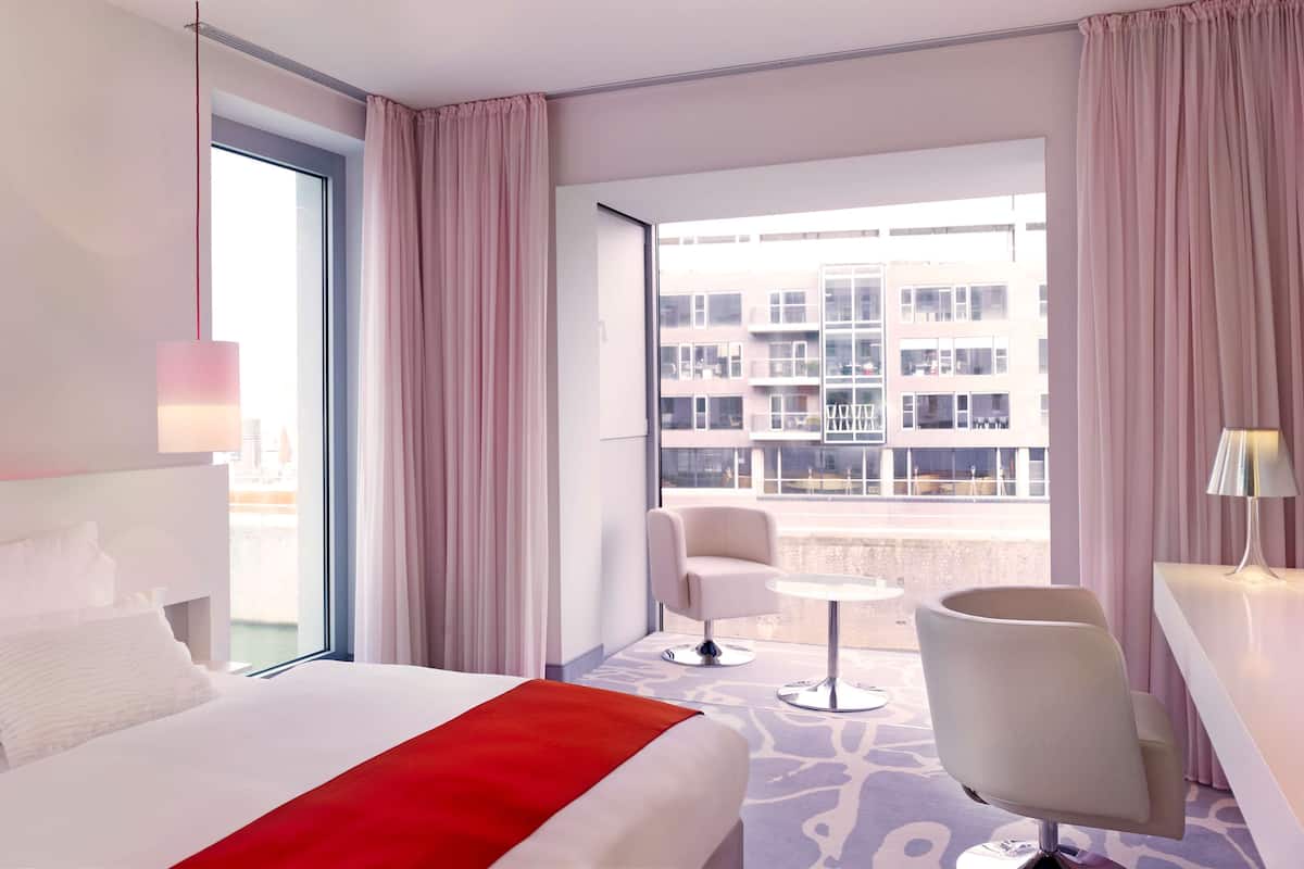 Ett hotellrum med en säng, en stol, ett litet bord och utsikt över en byggnad genom fönstret.