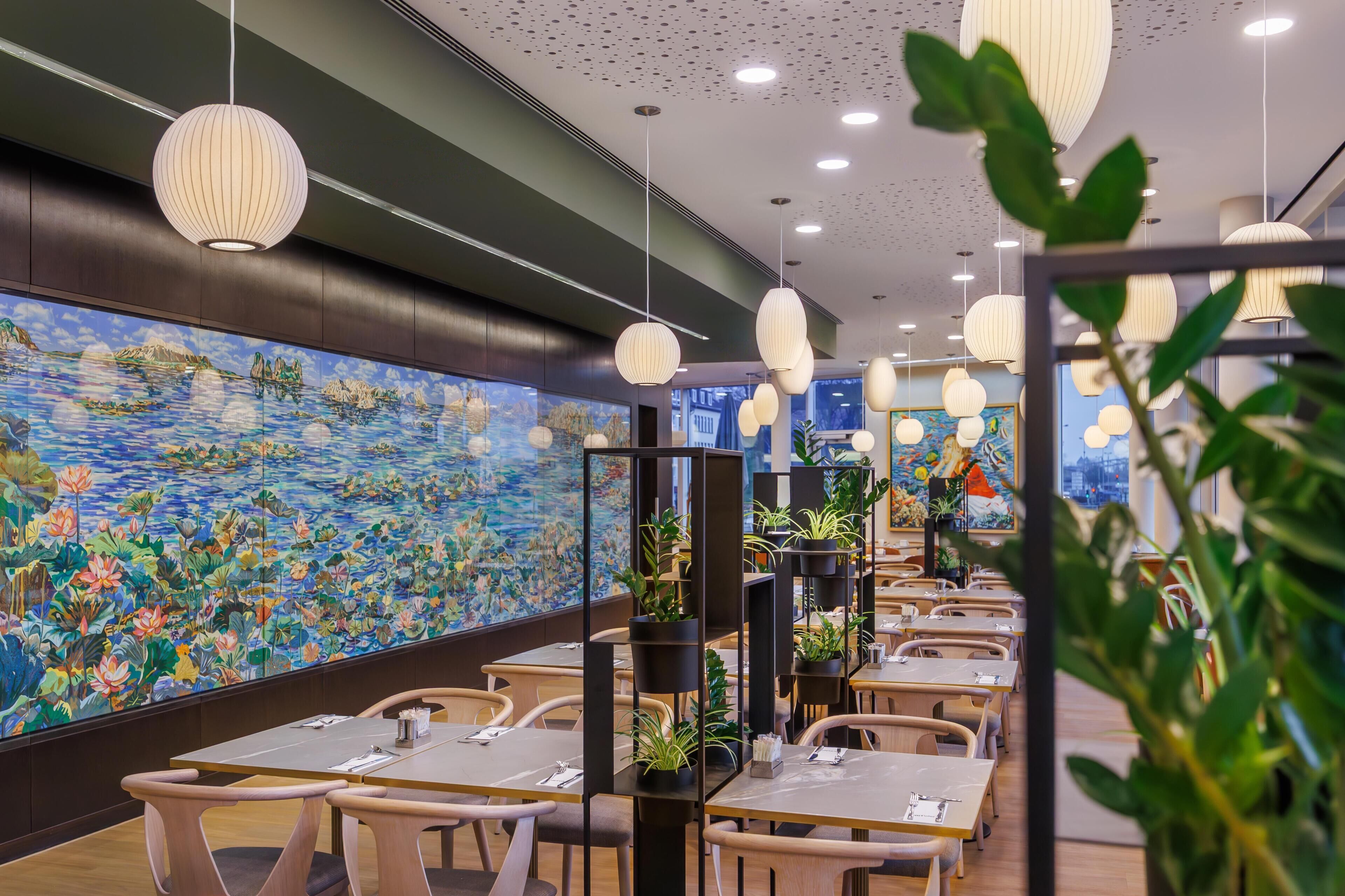 A restaurant with tables, chairs, hanging pendant lights, and a large mural of a landscape.
