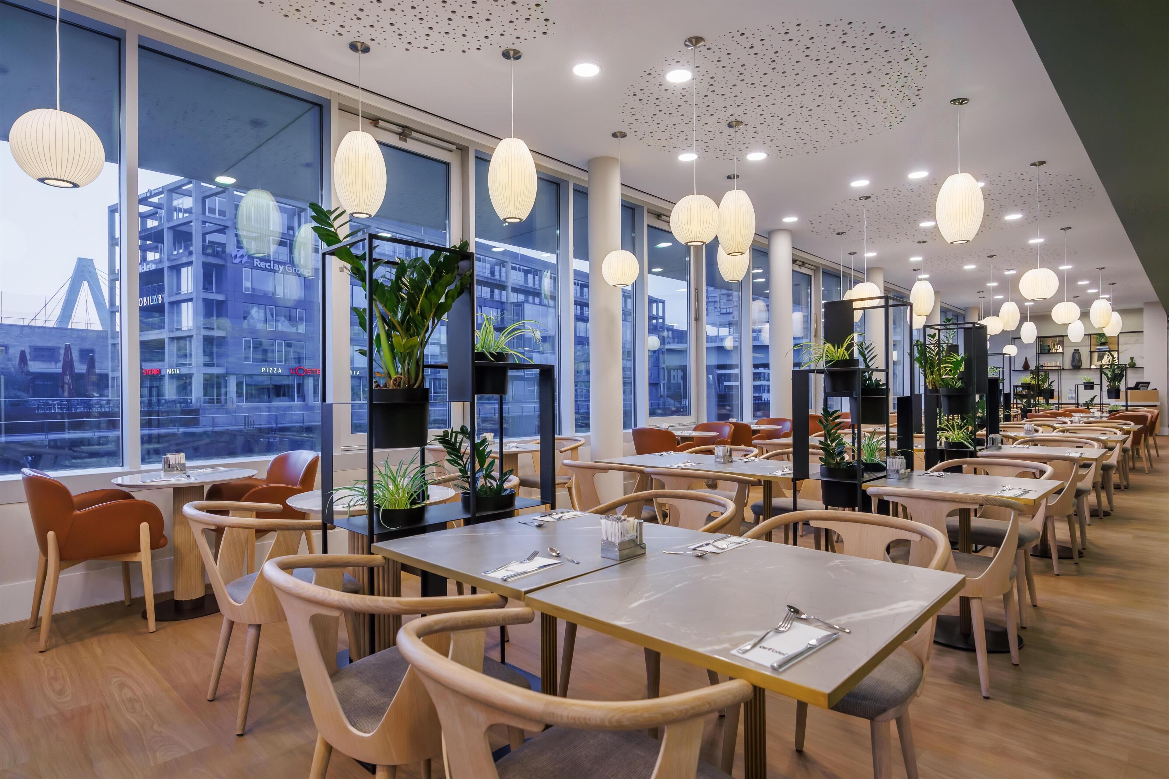 En modern restaurang med träbord och stolar, stora fönster och hängande taklampor.