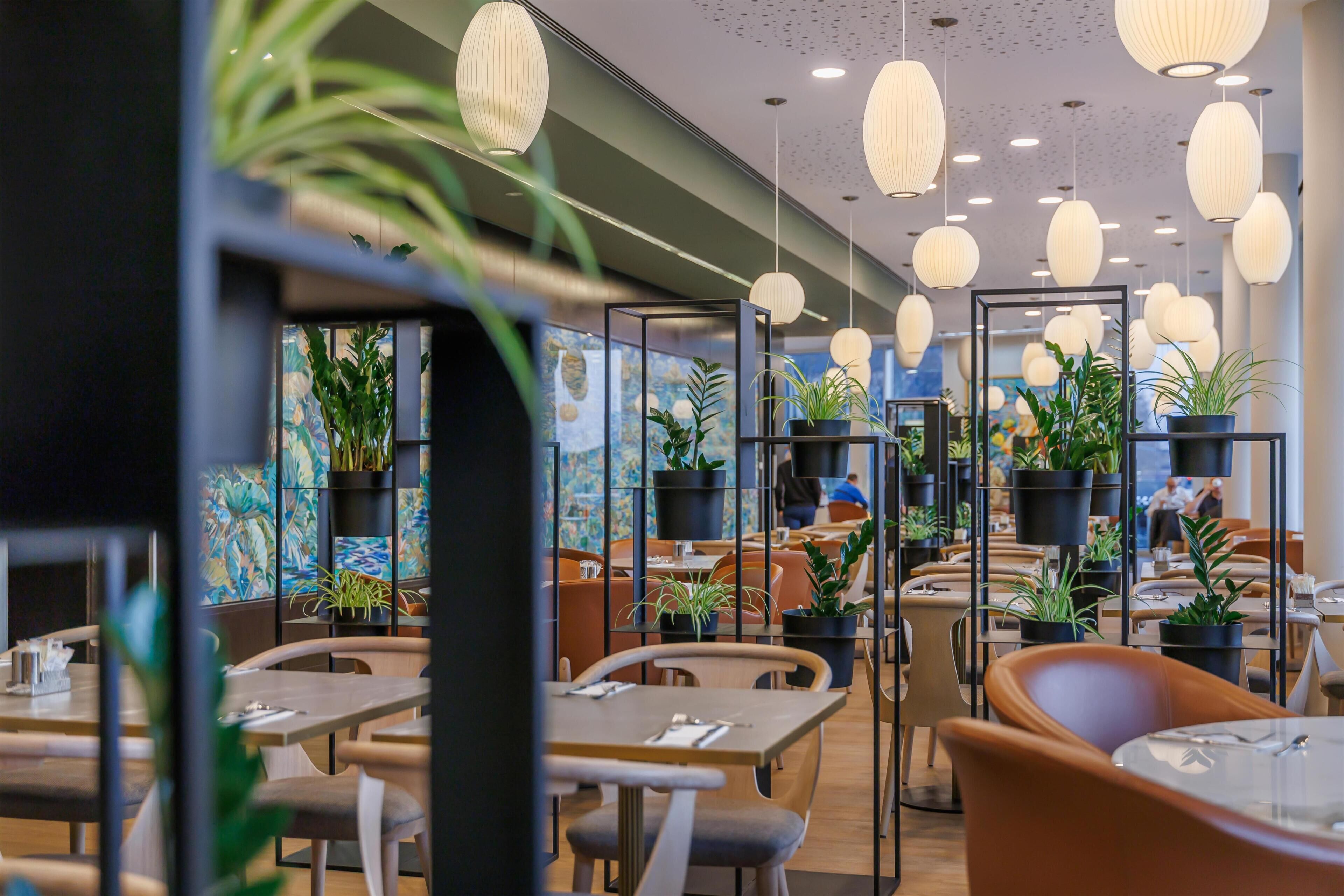 Un interior de restaurante moderno con mesas, sillas, plantas en macetas y lámparas colgantes.