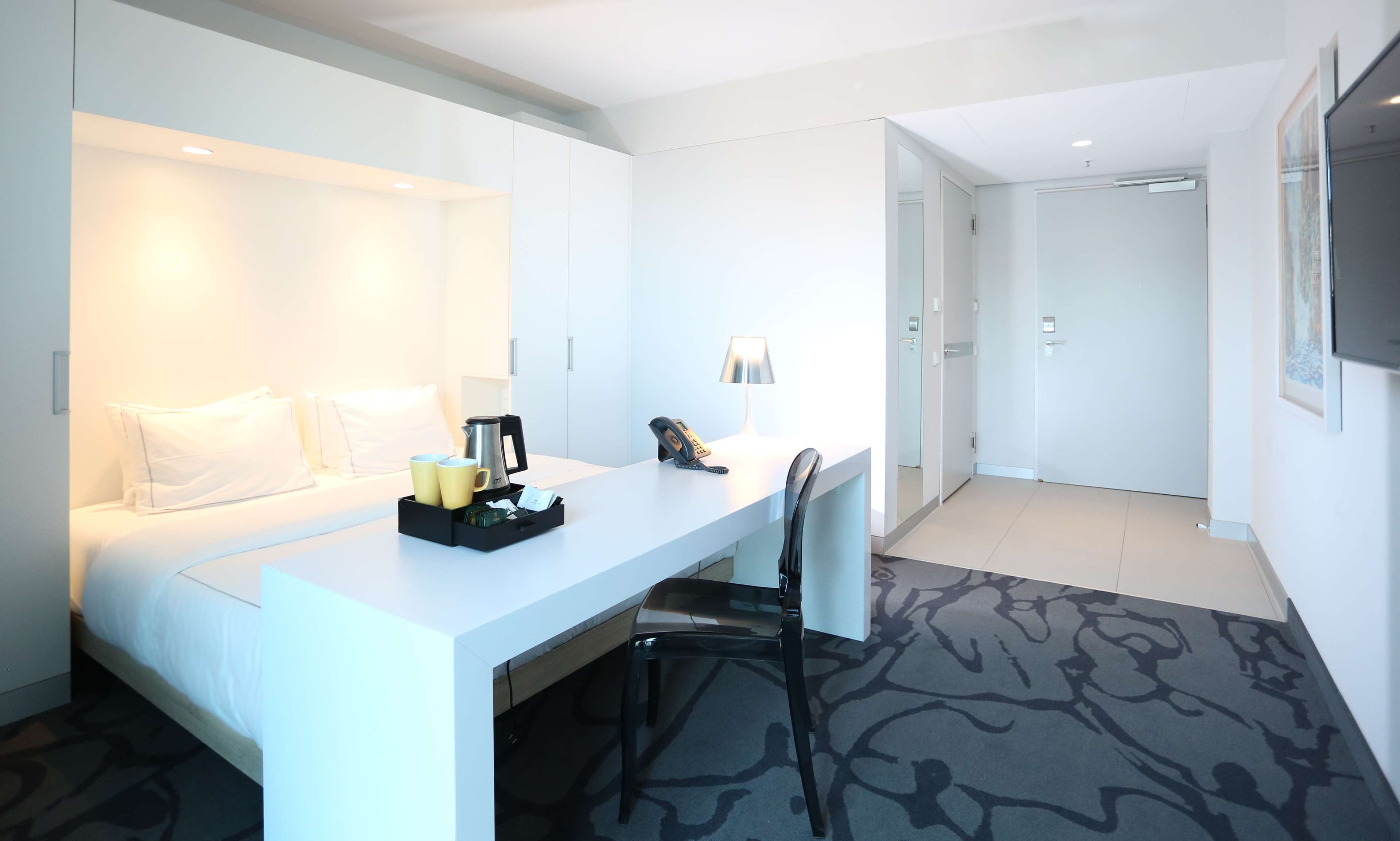 Ett hotellrum med en säng, ett skrivbord, en stol, en lampa och en kaffebryggare.