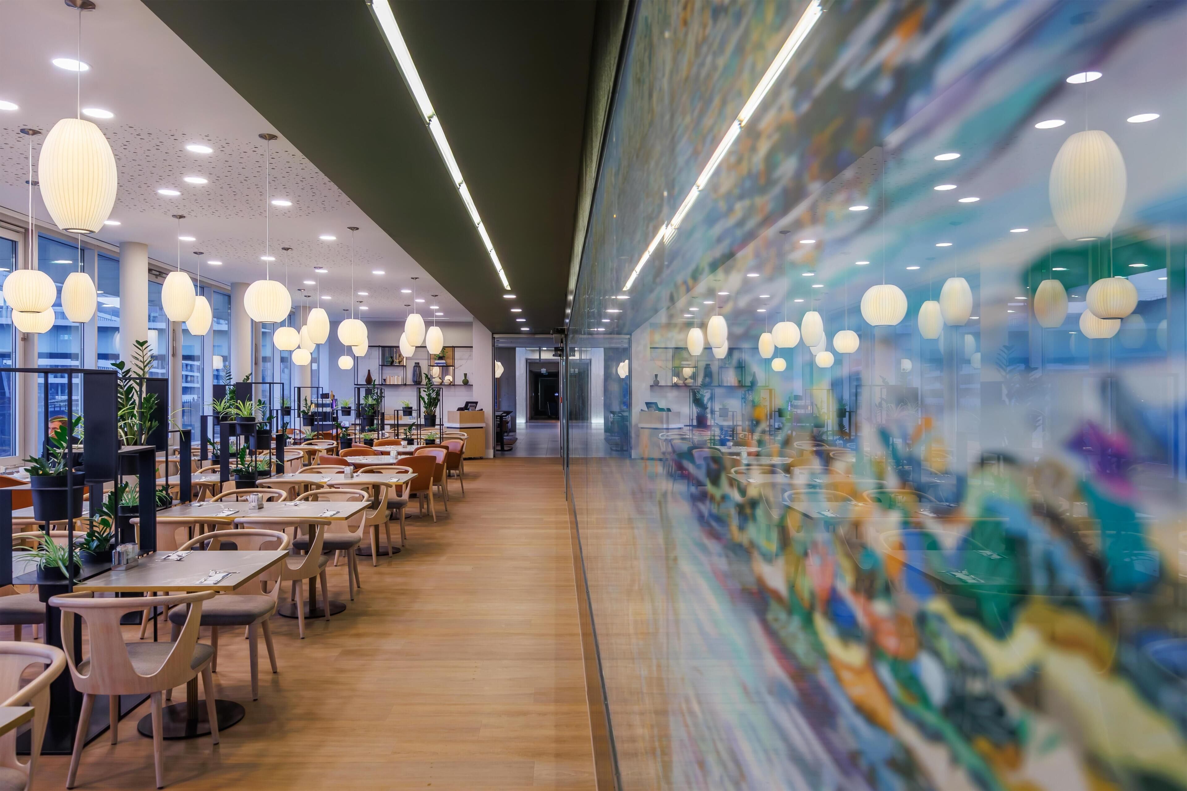 Un interior moderno con mesas y sillas, una pared de vidrio con obras de arte e iluminación colgante.