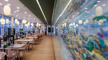 Ein modernes Interieur mit Tischen und Stühlen, einer Glaswand mit Kunstwerken und Pendelleuchten.