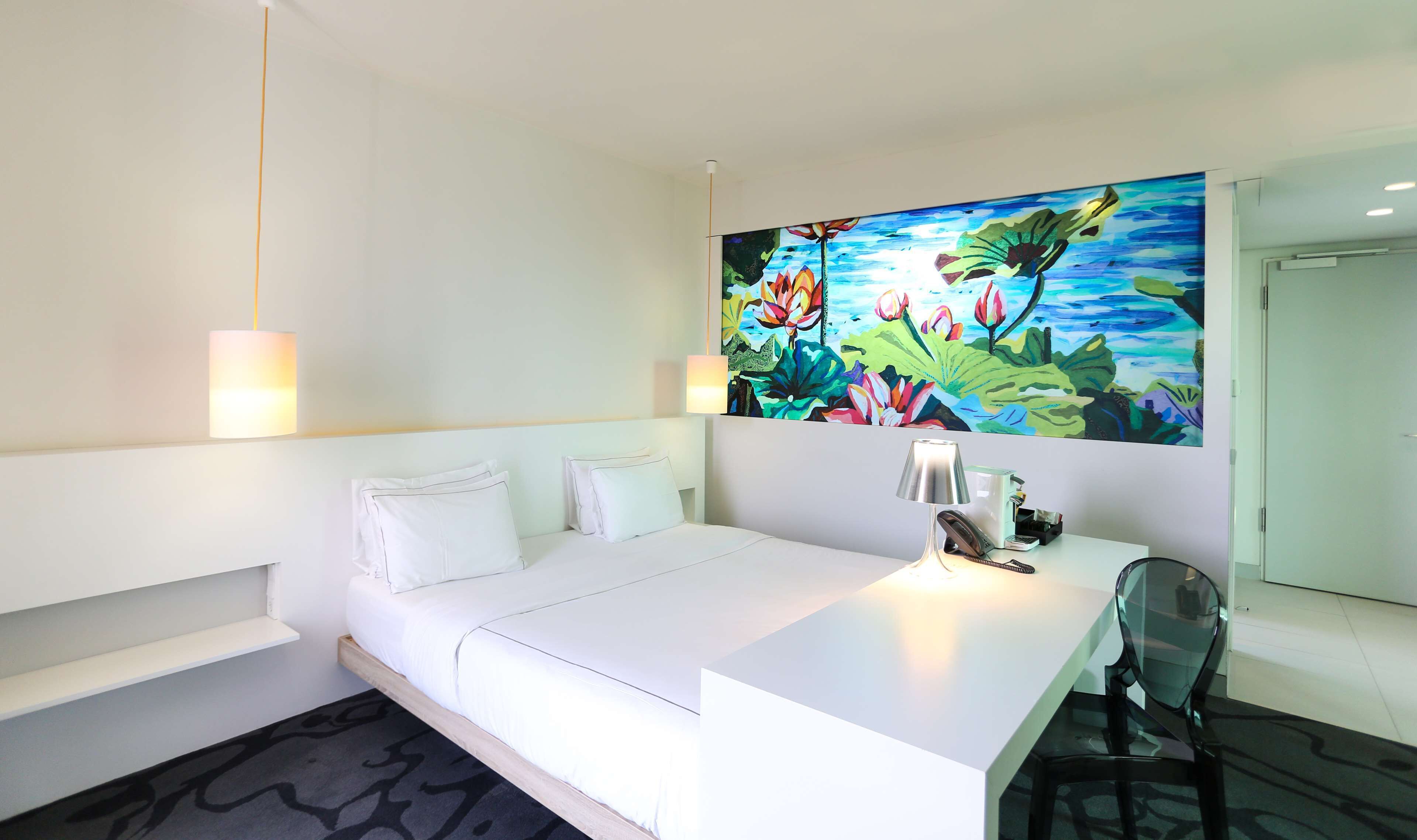 A hotel room with a bed, a desk, a chair, a painting, and a lamp.
