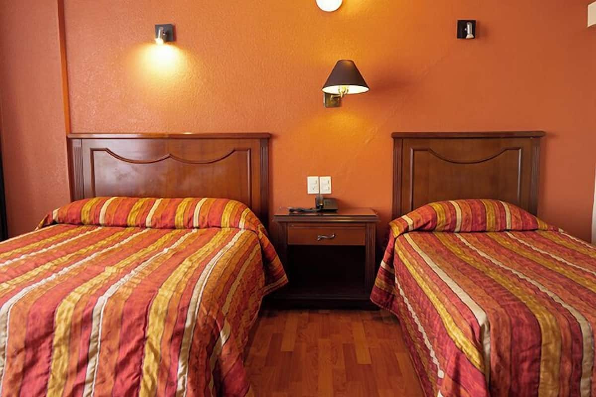 Una habitación con dos camas, cada una con ropa de cama a rayas, una mesita de noche con lámpara y un cajón pequeño.