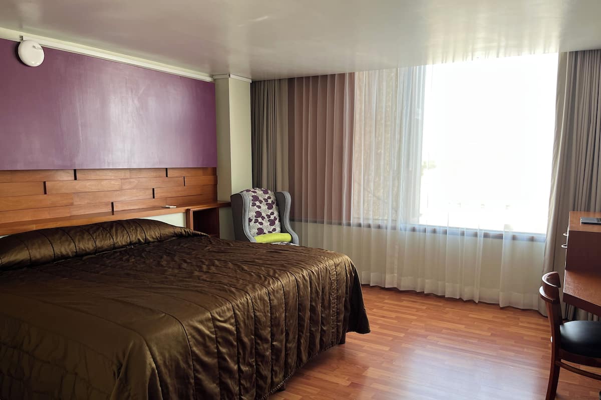 Una habitación de hotel con una cama grande, cabecera de madera, una silla, un escritorio y una ventana con cortinas.