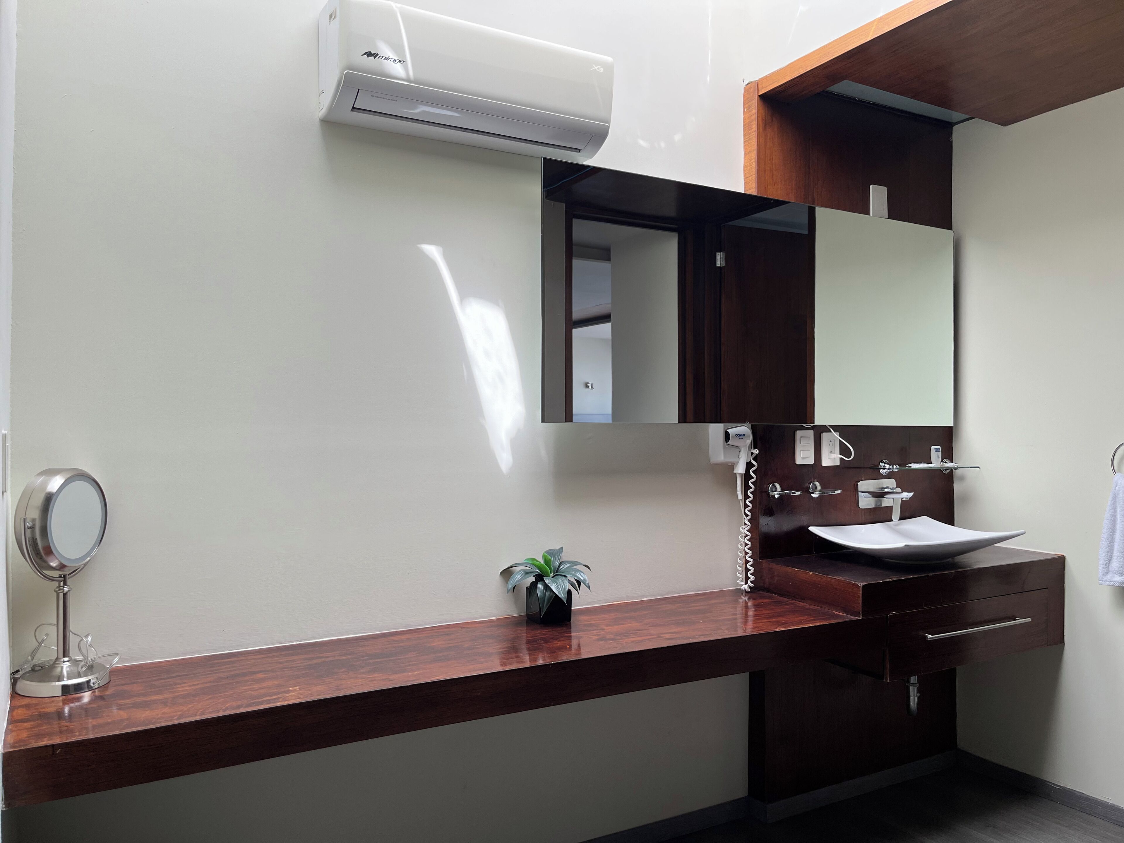 Un baño moderno con un tocador de madera, un lavabo blanco, un espejo y una unidad de aire acondicionado en la pared.