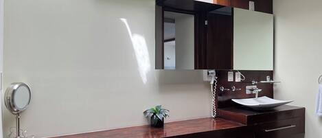Un baño moderno con un tocador de madera, un lavabo blanco, un espejo y una unidad de aire acondicionado en la pared.