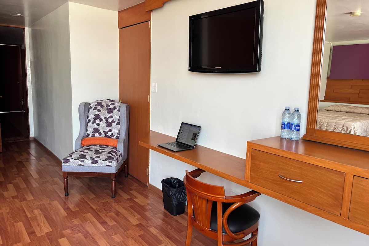 Habitación de hotel con un escritorio de madera, una laptop, una silla, un televisor montado en la pared y una cama con cabecera.