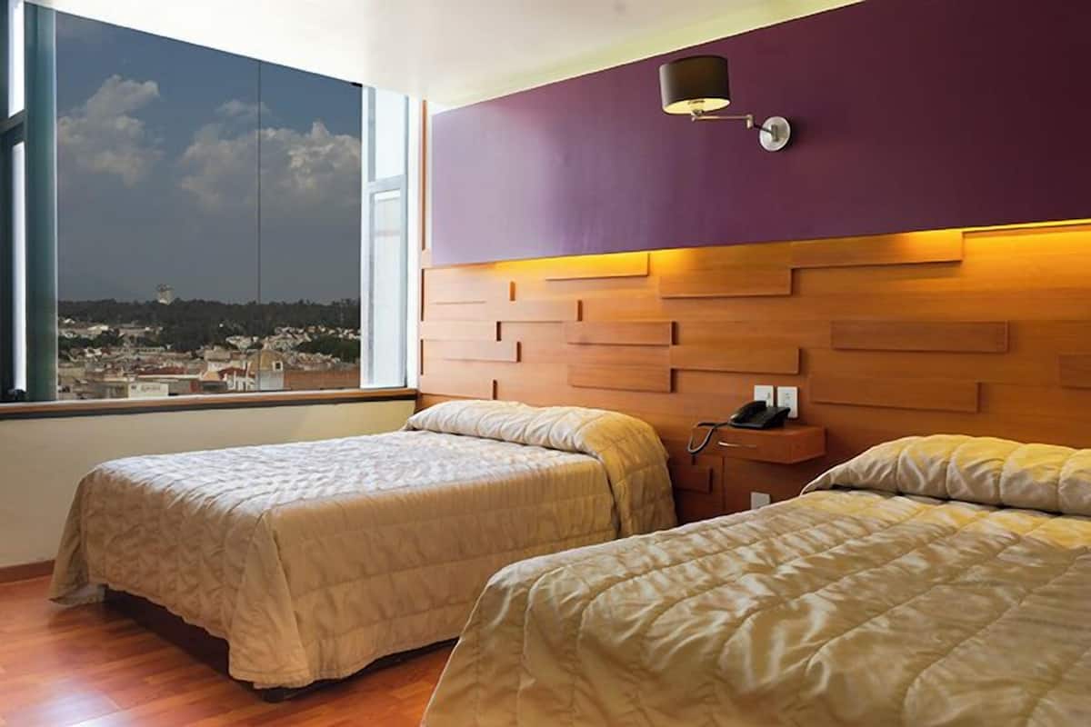 Habitación de hotel con dos camas, cabecero de madera, mesita de noche y vista a un paisaje urbano a través de un ventanal grande.