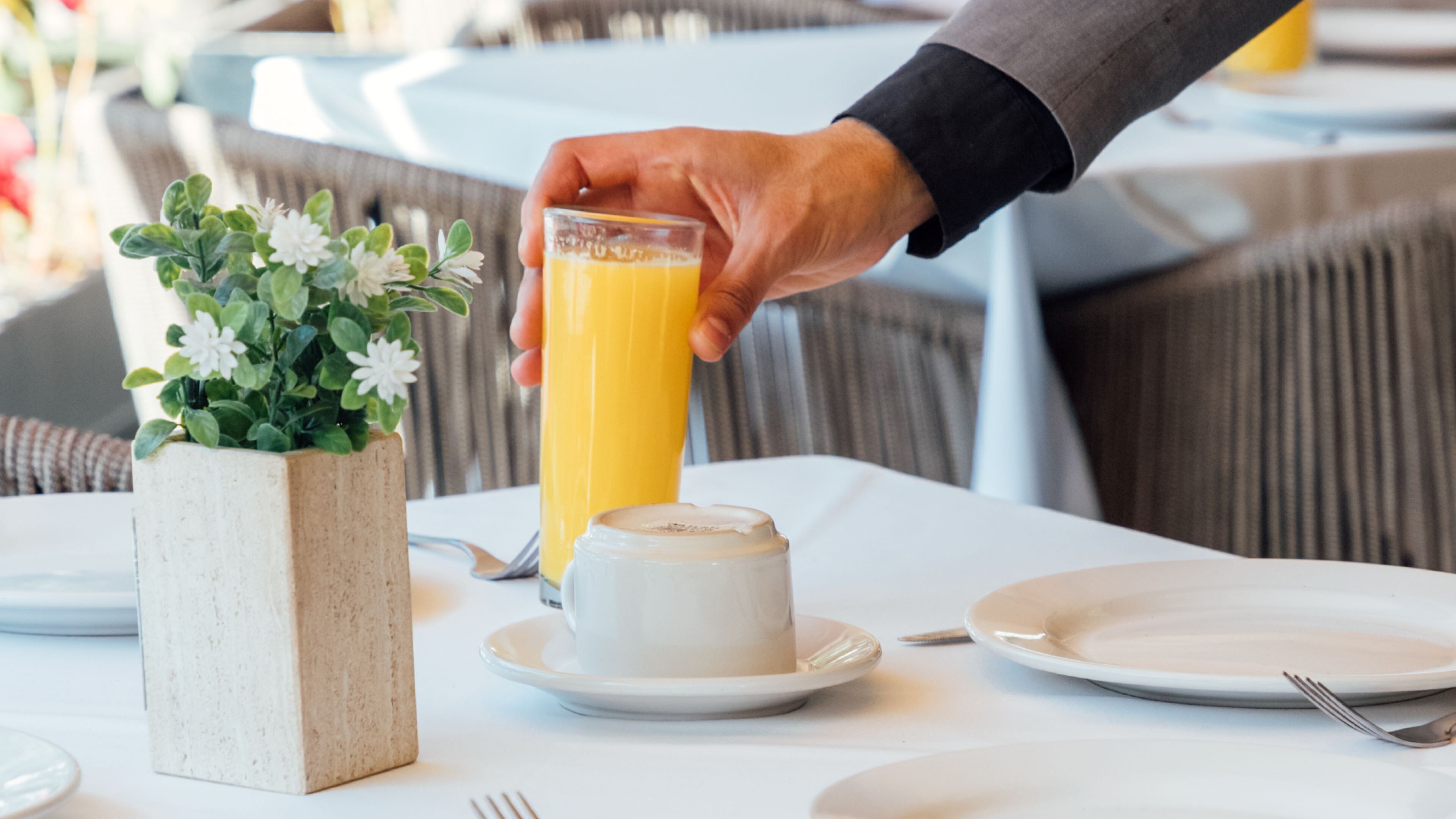 A dining table with a white plate, a glass of orange juice, a cup of coffee, and a small potted plant with white flowers.