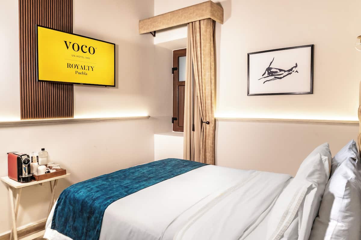 A hotel room with a bed, a bedside table with a camera and toiletries, a framed sign on the wall, and a door with curtains.