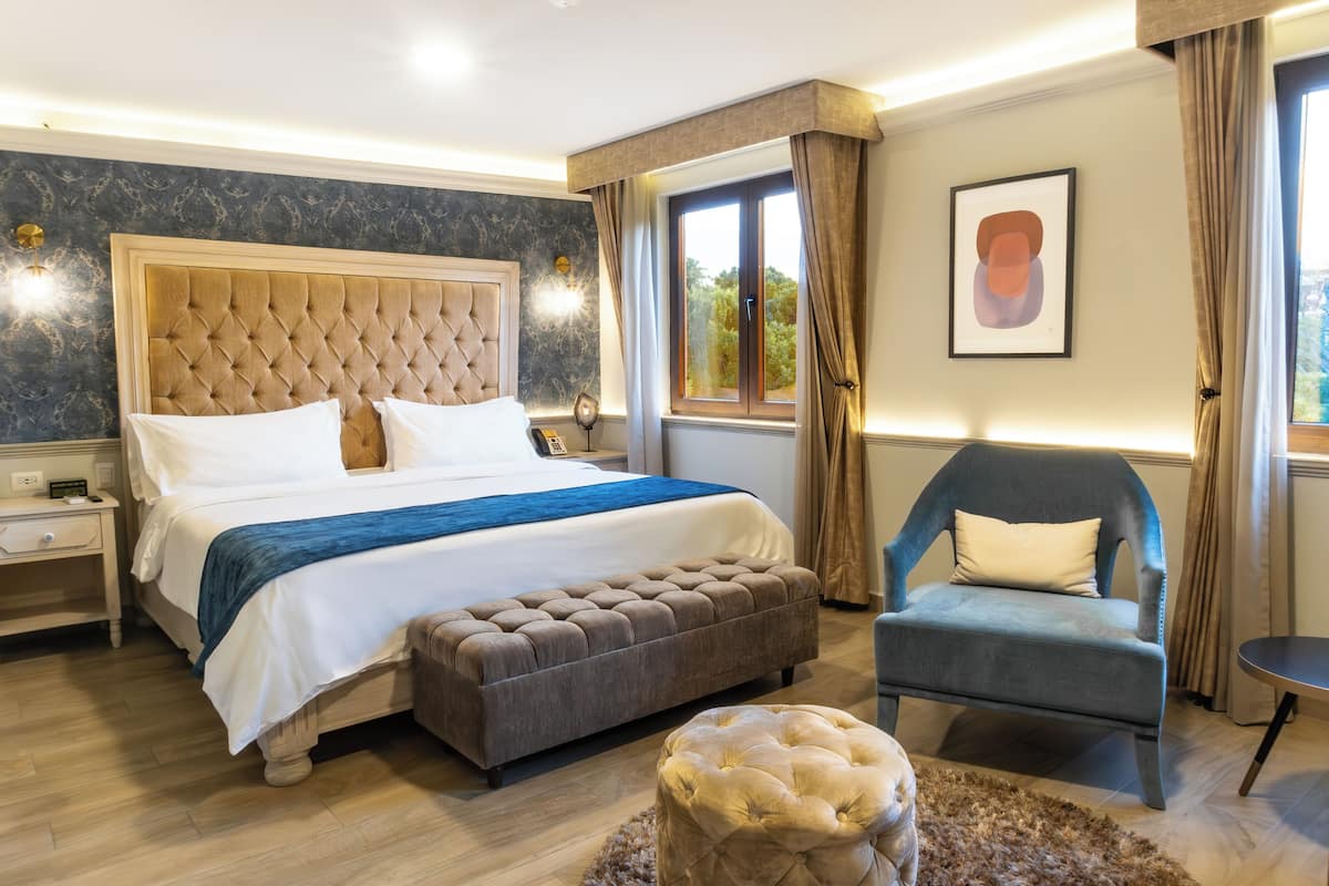A well-lit bedroom with a large bed, a tufted headboard, a bedside table, a chair, and a window with curtains.
