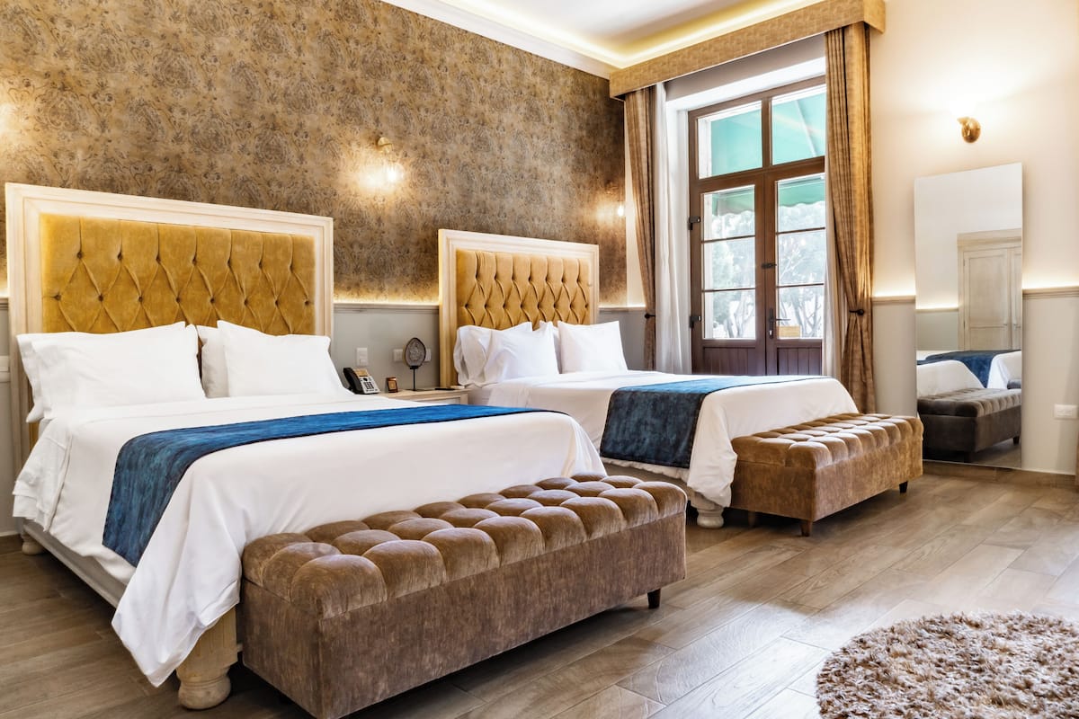 A hotel room with two beds, a large window with curtains, a wooden floor, and a mirror on the wall.
