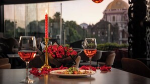 Une table dressée avec une bougie, des verres à vin et un vase de roses rouges, avec vue sur un bâtiment par la fenêtre.