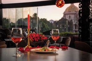 Une table dressée avec une bougie, des verres à vin et un vase de roses rouges, avec vue sur un bâtiment par la fenêtre.