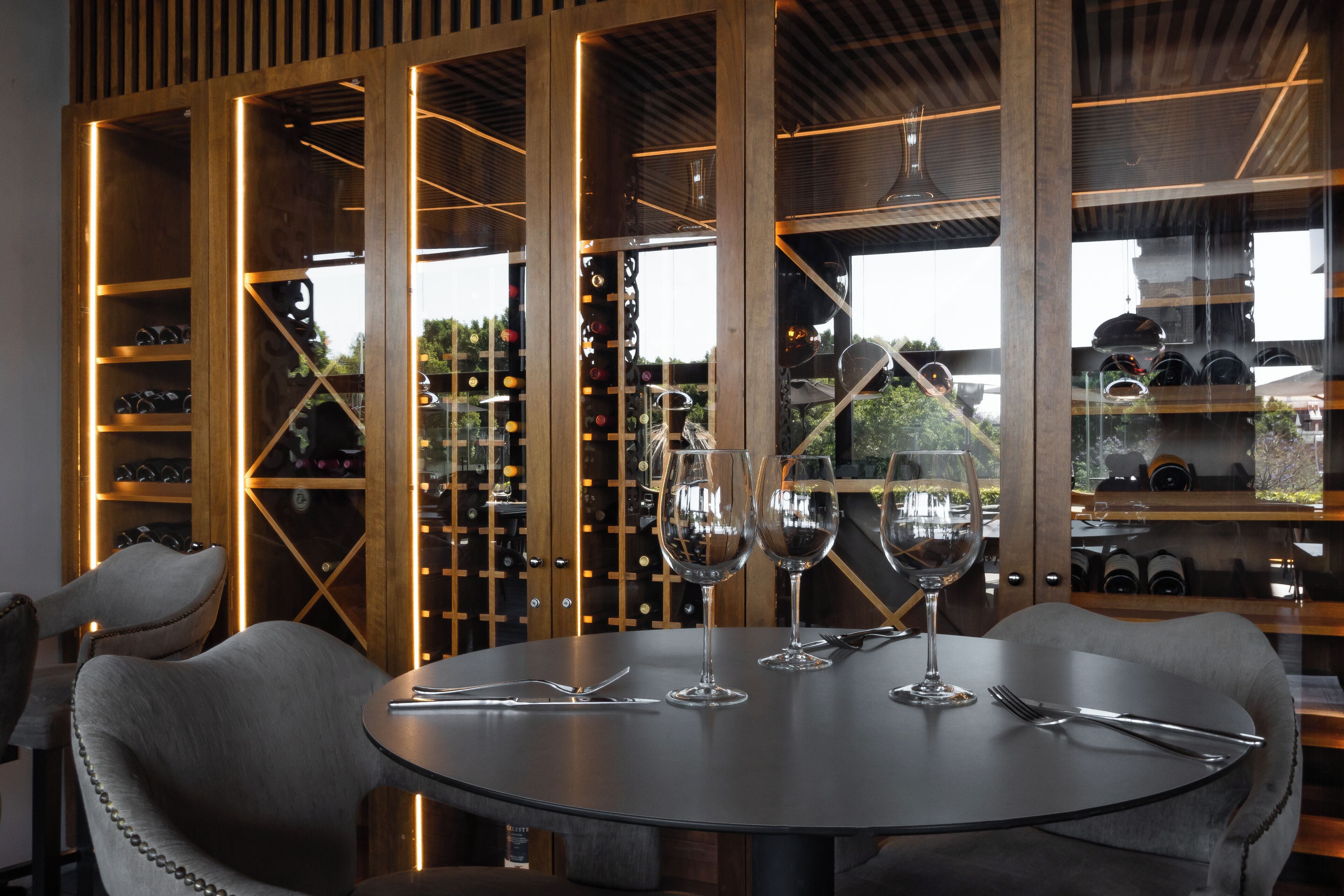 Une table ronde, agrémentée de verres à vin et de couverts, est placée devant un décor représentant une cave à vin avec des étagères en bois et des bouteilles.
