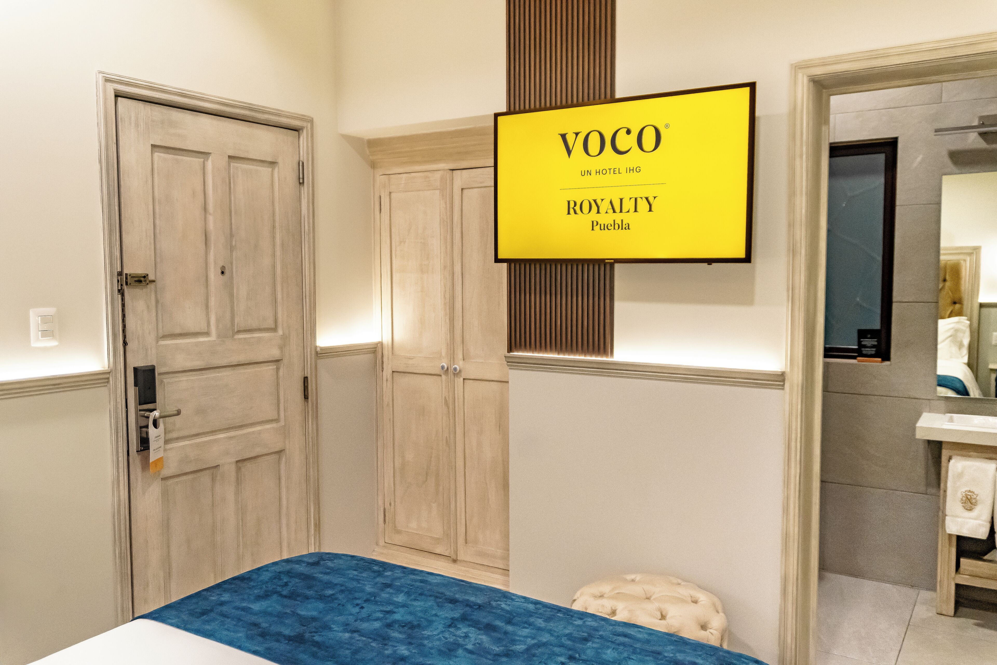A hotel room with a bed, a wardrobe, a vanity with a mirror, and a sign indicating the hotel name and location.