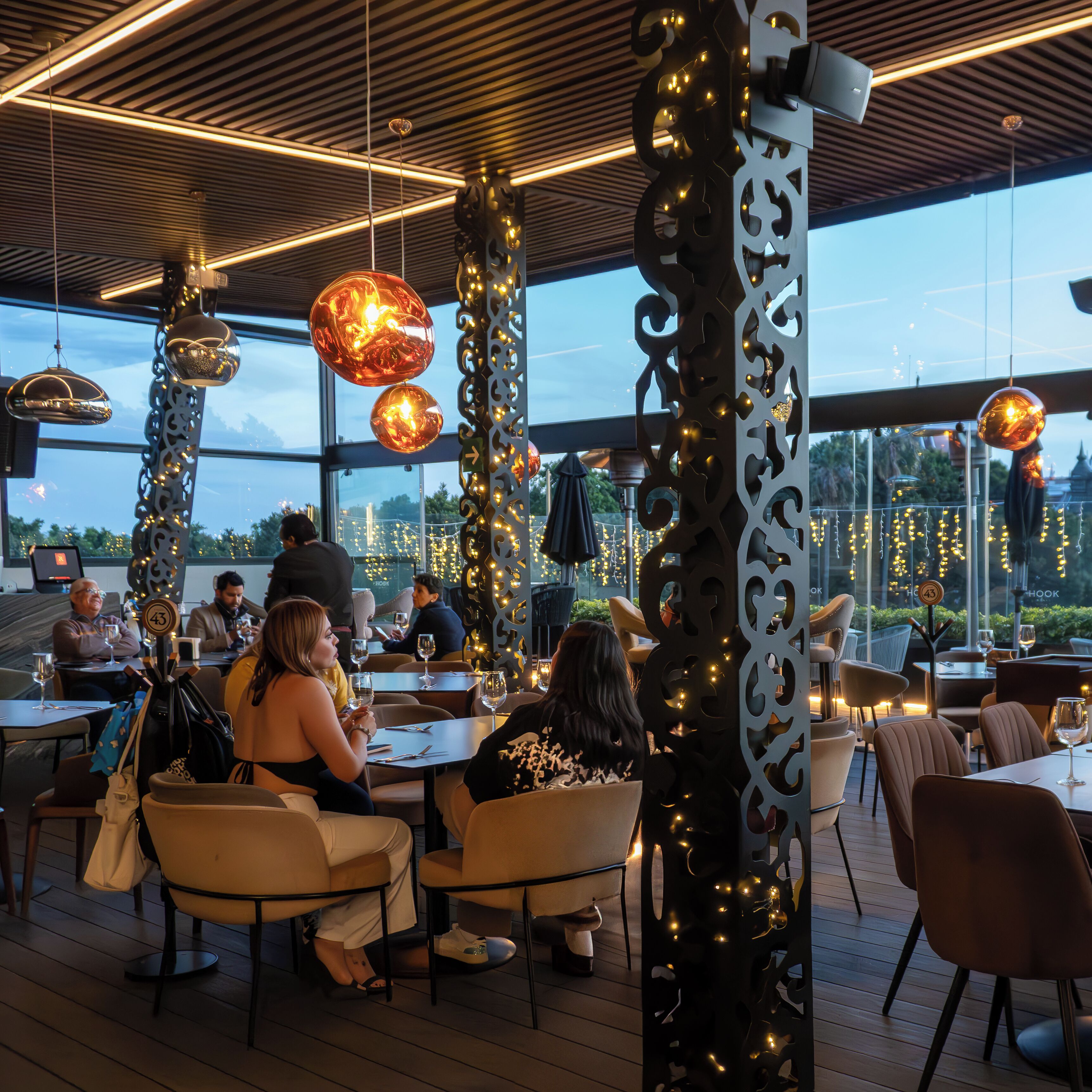 An indoor dining area with tables and chairs, decorative lighting, and large windows.