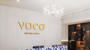 Un espace d’accueil agrémenté d’un lustre, d’un comptoir de réception et d’un mur sur lequel est inscrit le nom de l’hôtel : « Voco » et « Un hôtel IHG ».