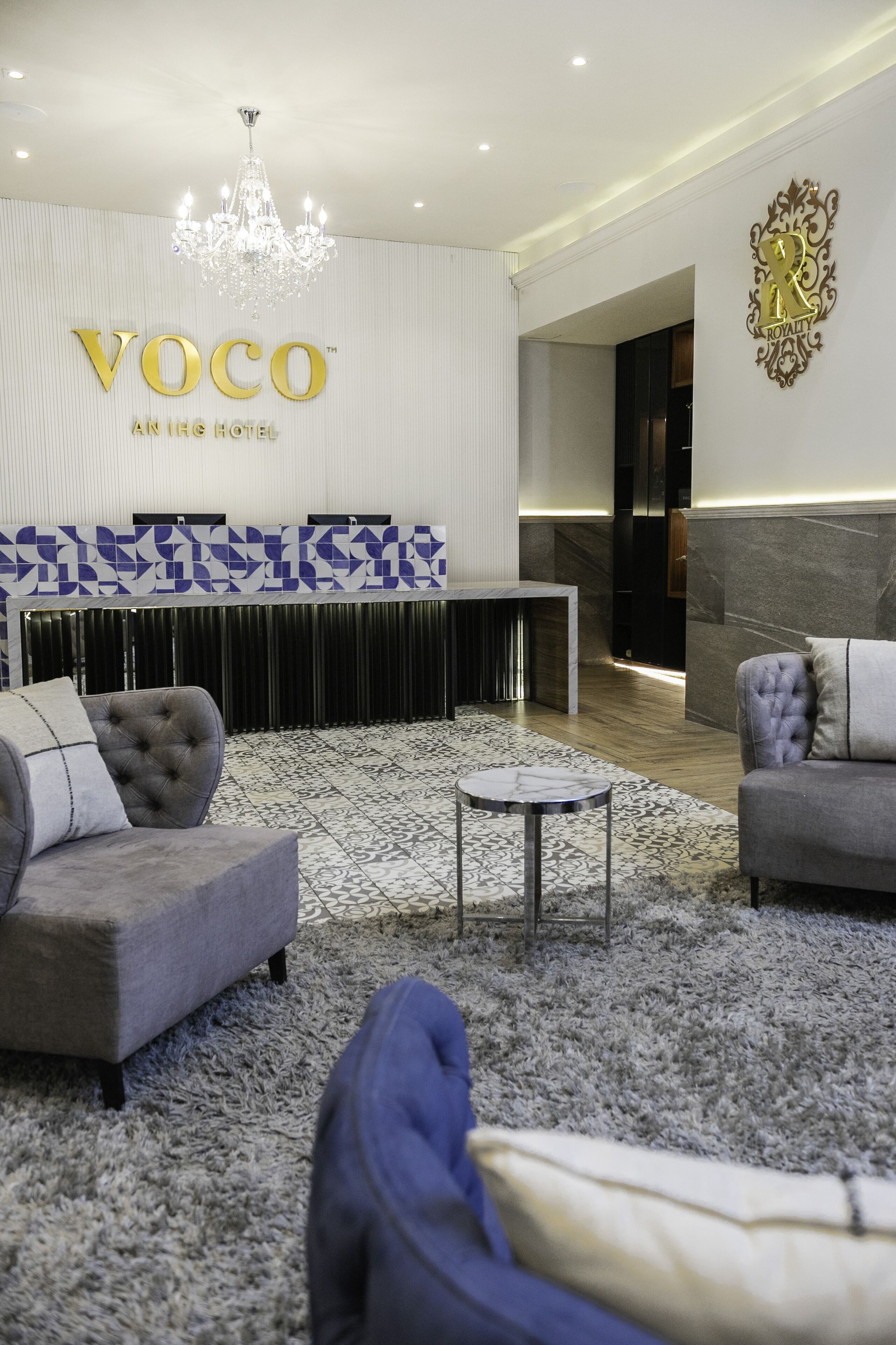 A modern hotel lobby with a chandelier, a reception desk, and a seating area with sofas and a coffee table.