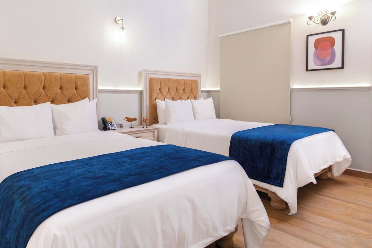 A hotel room with two beds, each with a tufted headboard and white bedding. There is a small bedside table with a telephone and a decorative bird. The room has a light-colored wall and a framed picture above the beds.