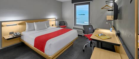 A hotel room with a bed, desk, chair, and a window with a view of the sea.
