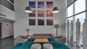 Un área de descanso moderna con un sofá gris, dos otomanas, una mesa de centro de madera y un tapete color turquesa. Seis pinturas de atardecer adornan la pared, junto con dos lámparas de pie y altas columnas de concreto.