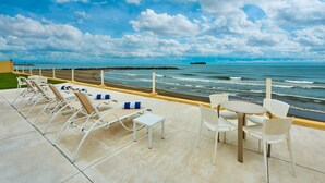 Una terraza frente al mar con sillones y mesas, con vistas al océano y un rompeolas rocoso.
