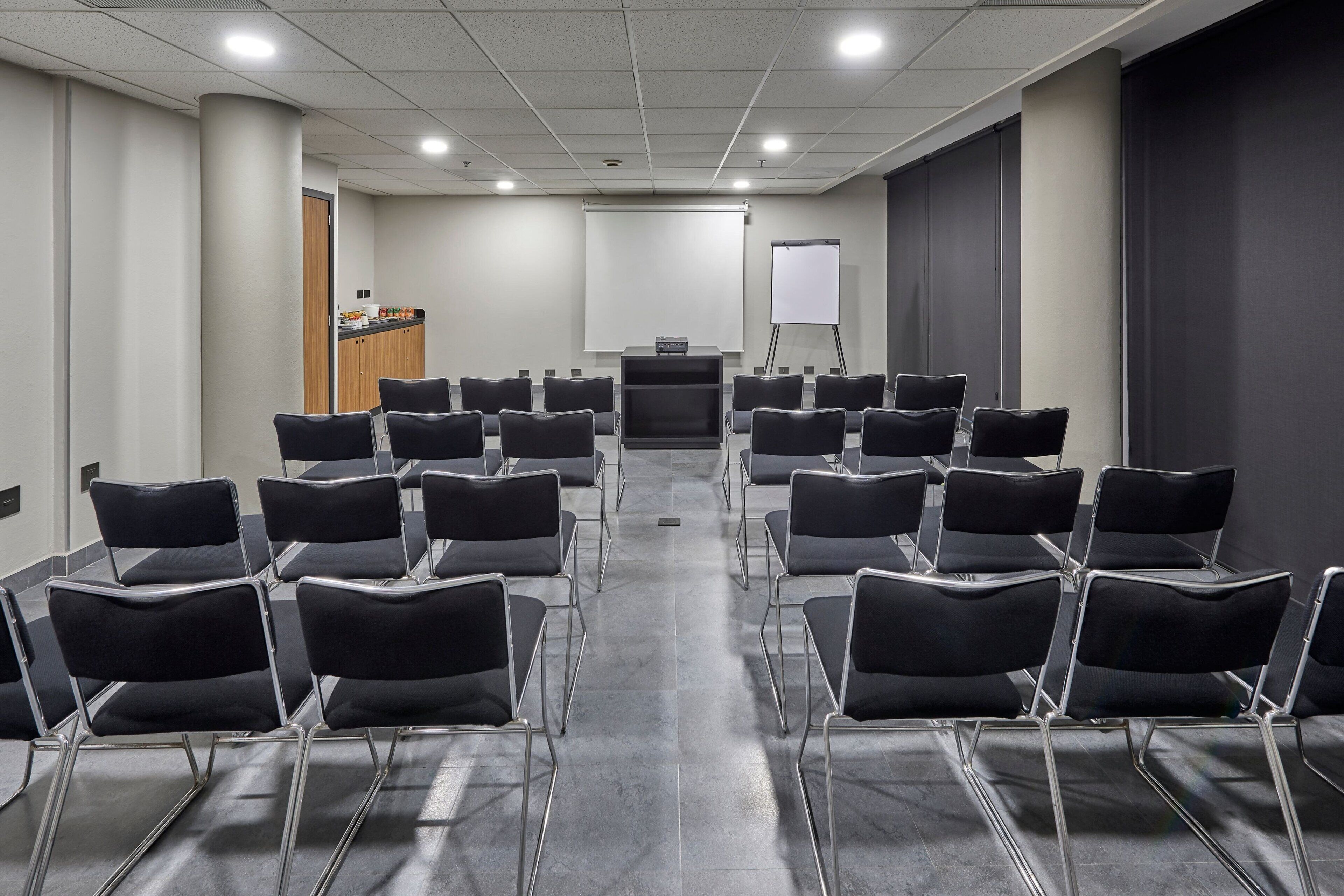 A conference room with rows of black chairs, a podium, a whiteboard, and a projector screen.