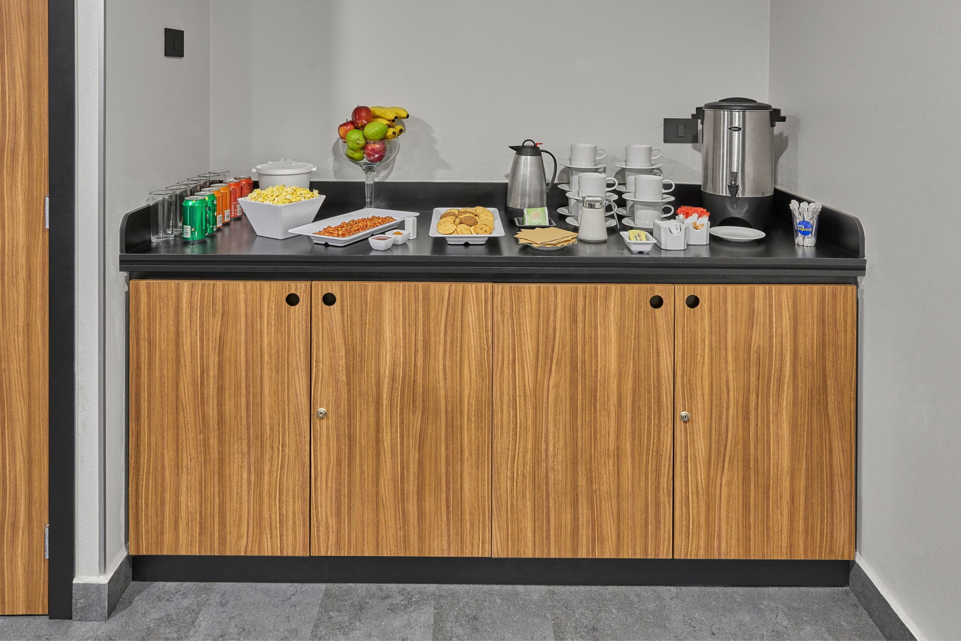 A kitchenette with a black countertop, wooden cabinets, and various food and drink items.