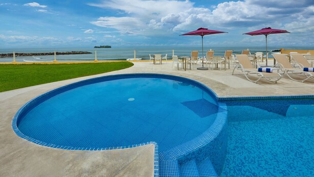 Una piscina con sillas de descanso y sombrillas, un área verde y vistas al océano.