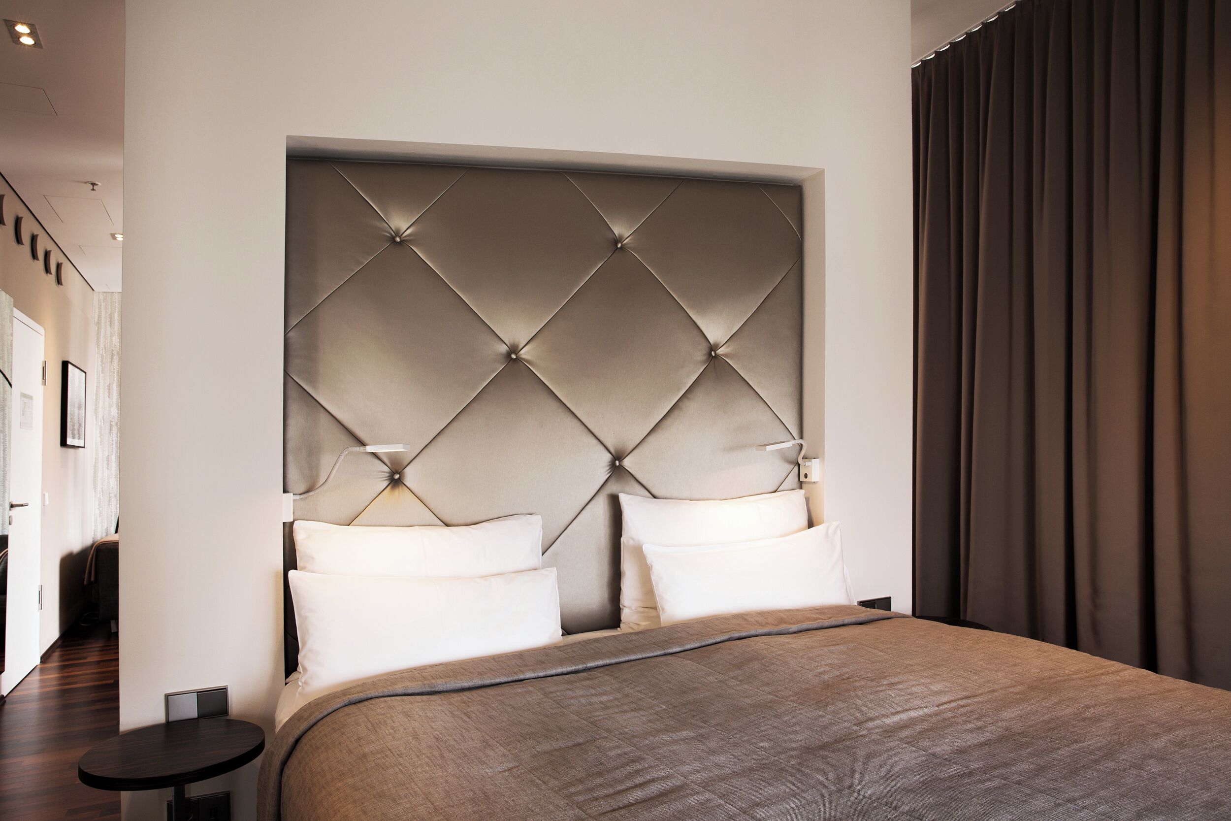 A modern hotel room with a large bed, a tufted headboard, and a dark curtain.