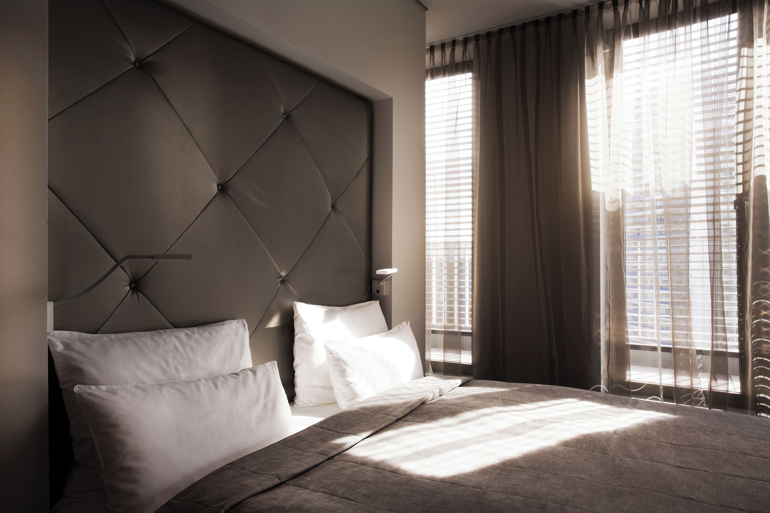 A bedroom with a tufted headboard, a large bed with white linens, and a view through sheer curtains to the outside.