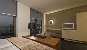 Ein modernes Hotelzimmer mit einem großen Bett, einem Schreibtisch mit Lampe, einem Fernseher und Blick auf die beleuchtete Stadt bei Nacht.