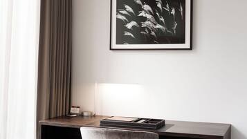 A hotel room with a desk, a chair, a framed picture, and a lamp.