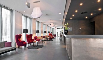 Eine moderne Hotelhalle mit roten Stühlen, einem Empfangsbereich und großen Fenstern mit leichten Vorhängen.