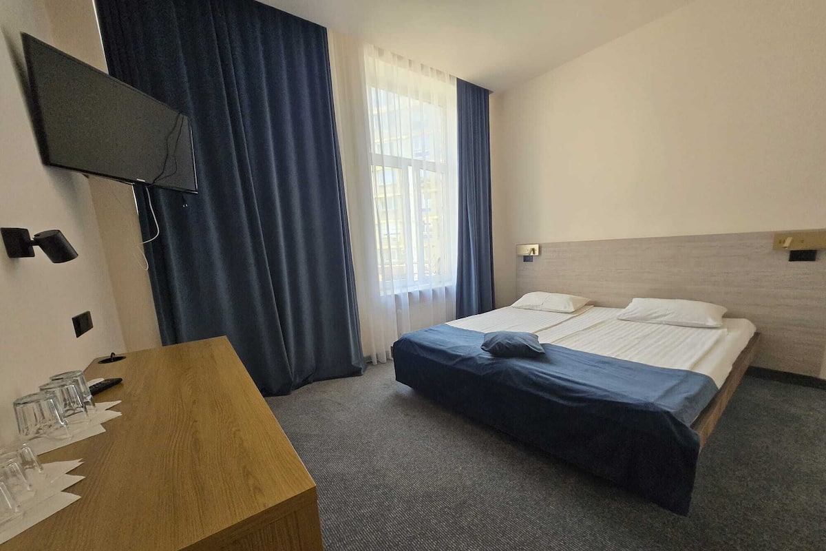 Habitación de hotel con cama, escritorio con vasos, televisión y ventana con cortinas.
