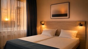 Una cama doble con dos almohadas, un cabecero con luces empotradas en la pared, una ventana con cortinas traslúcidas y un cuadro enmarcado en la pared.