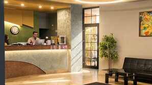 Zona de recepción de hotel con recepcionista, reloj, mostrador de recepción, máquina de café, planta, sofá y puerta que da al exterior.