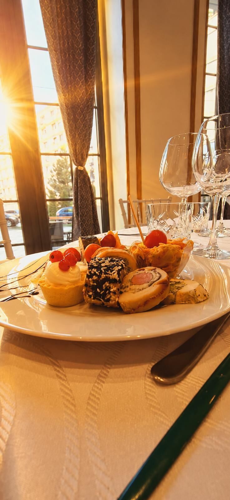 Une assiette de desserts est posée sur une table, à côté de verres  à vin et d’une fourchette, près d’une fenêtre ornée de rideaux.