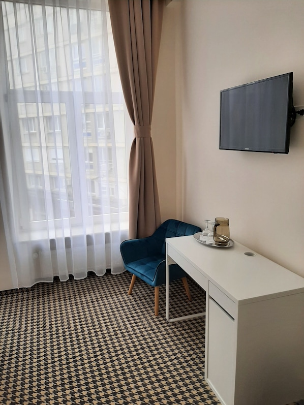 A room with a patterned floor, a desk with a chair, a television on the wall, and a window with curtains.