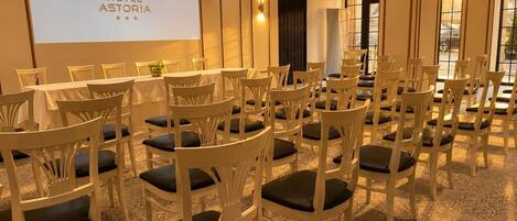 Ein Konferenzraum mit mehreren weißen Stühlen und einer Projektionsleinwand, die 'HOTEL ASTORIA' zeigt.