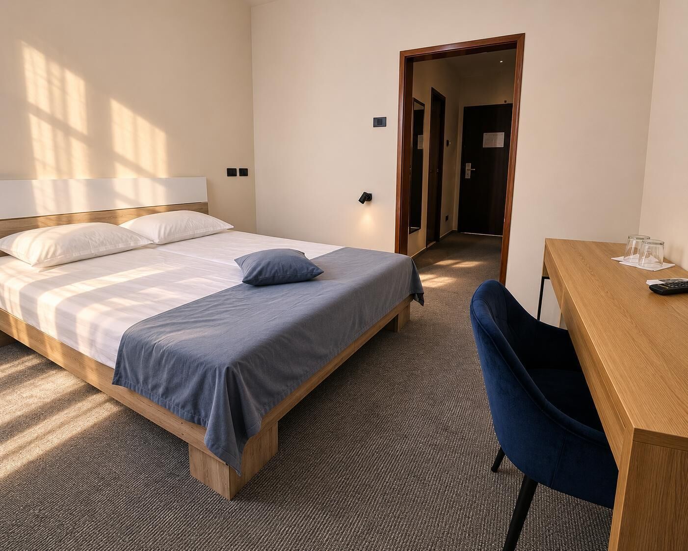 A hotel room with a bed, a desk, and a chair. There is a door leading to another room.