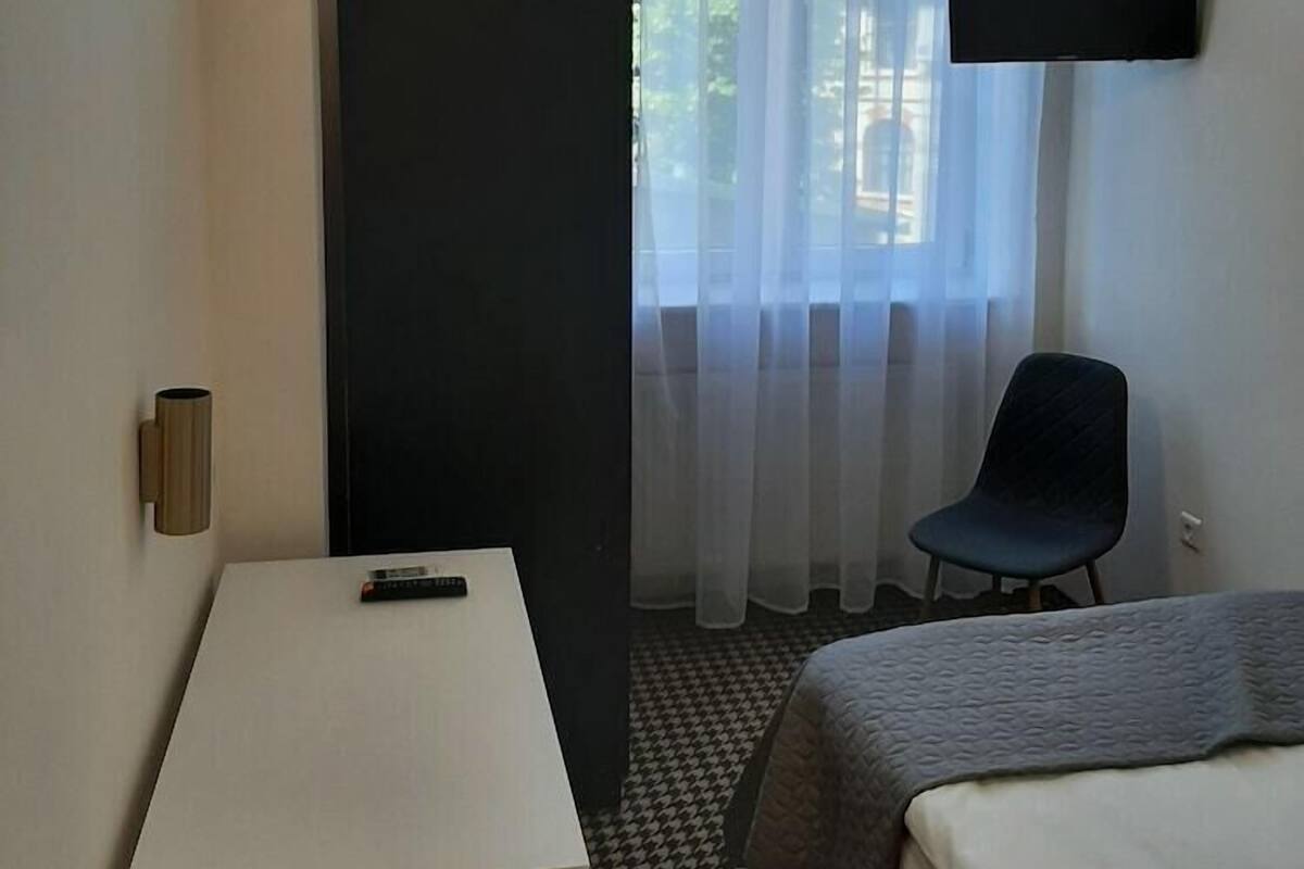 Una habitación de hotel con cama, escritorio, silla, televisor montado en la pared y ventana con cortinas traslúcidas.