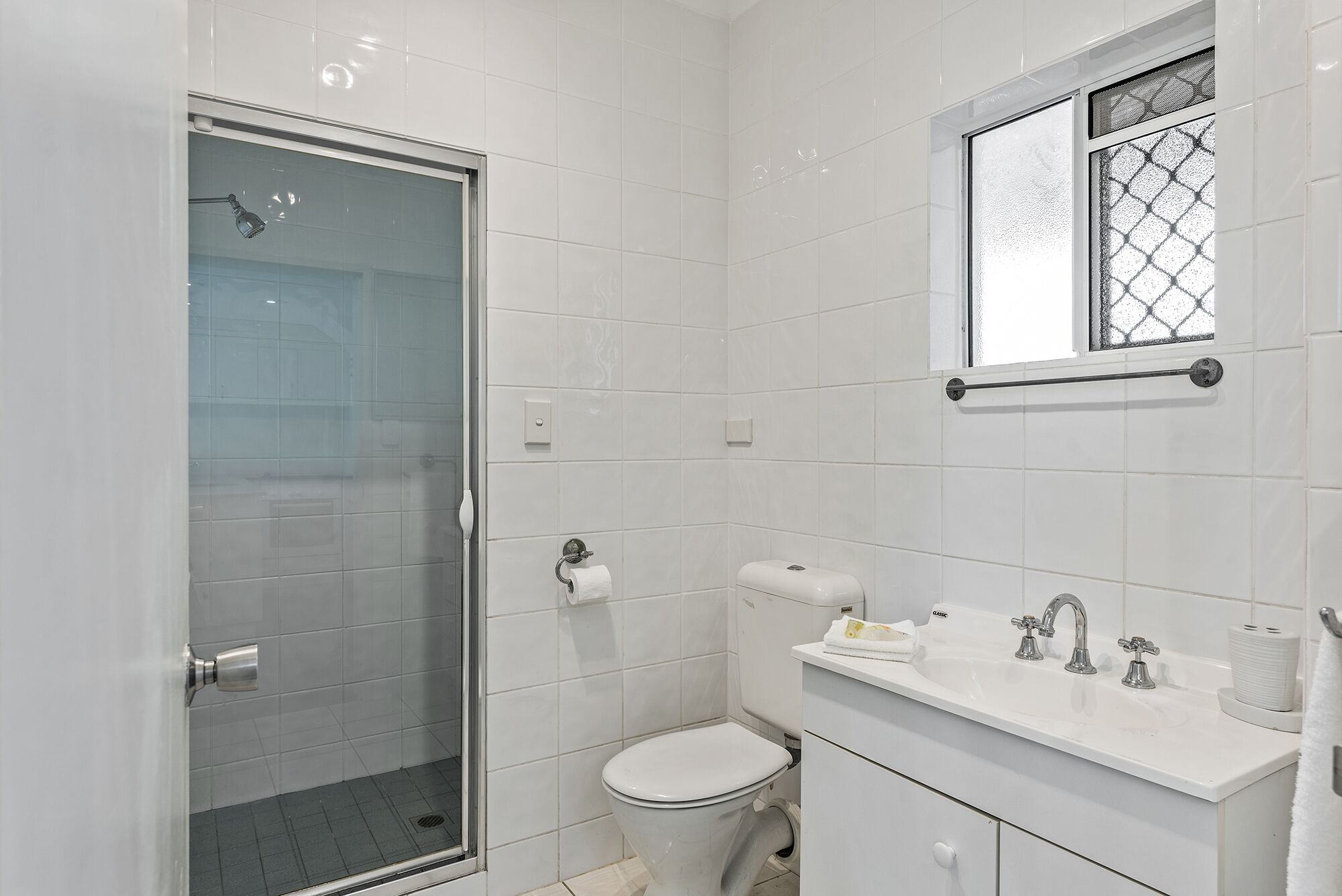 Une salle de bain avec une douche à parois de verre, des toilettes, un lavabo avec un miroir et une fenêtre ornée d’une grille décorative.