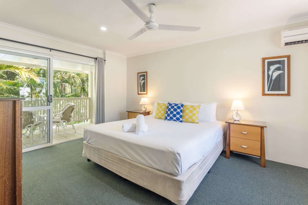 Una habitación de hotel con cama, dos mesitas de noche, ventilador de techo y balcón con mobiliario exterior.