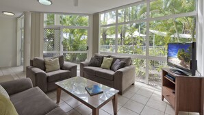 A spacious living room with two sofas, a coffee table, and a TV on a wooden stand. Large windows offer a view of palm trees and a fence.