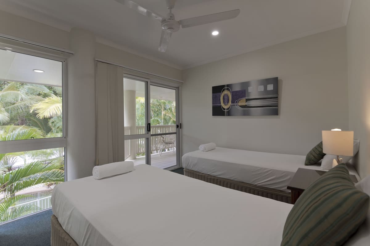 Habitación de hotel con dos camas, ventilador de techo, ventana con vista a palmeras y un cuadro en la pared.